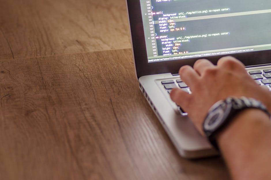 A person using a laptop on a wooden desk, with visible code on the screen, possibly related to gardening or landscaping management. The person's right hand, wearing a watch, is positioned over the key